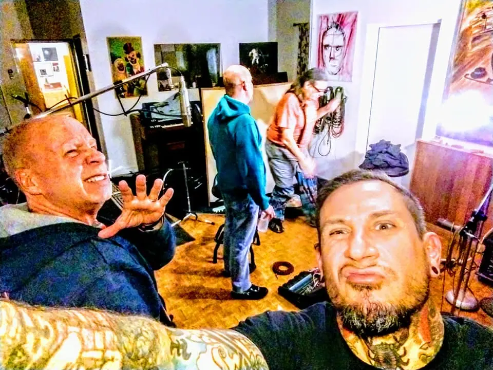 Goofy selfie in the live room: a tattooed man pulling a face in the foreground, an older man grimacing with his hand up, and two musicians setting up behind them under a clown painting.