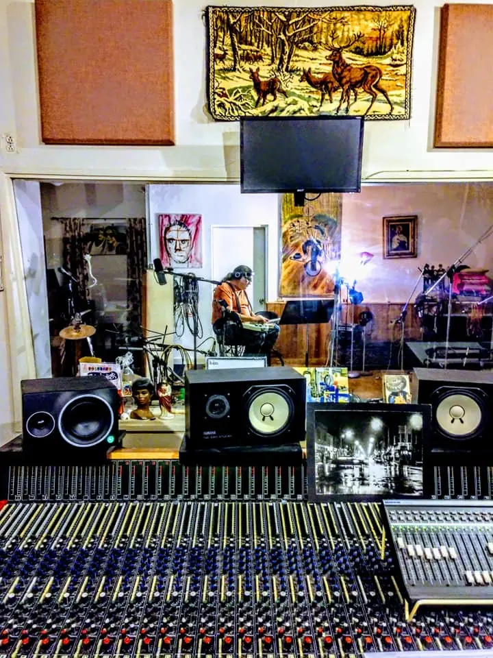 View over the console and Yamaha monitors through the glass into the live room, under a woven tapestry of deer in the snow.
