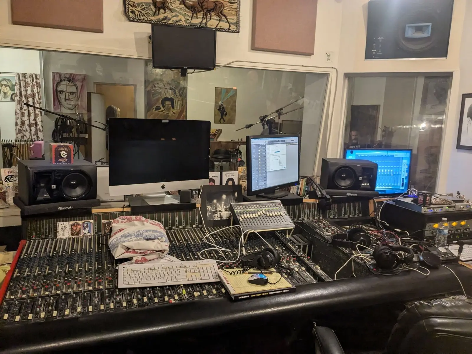 The control room mid-session: a long analog console, monitors, headphones, a keyboard and cables everywhere, with the live room visible through the glass.