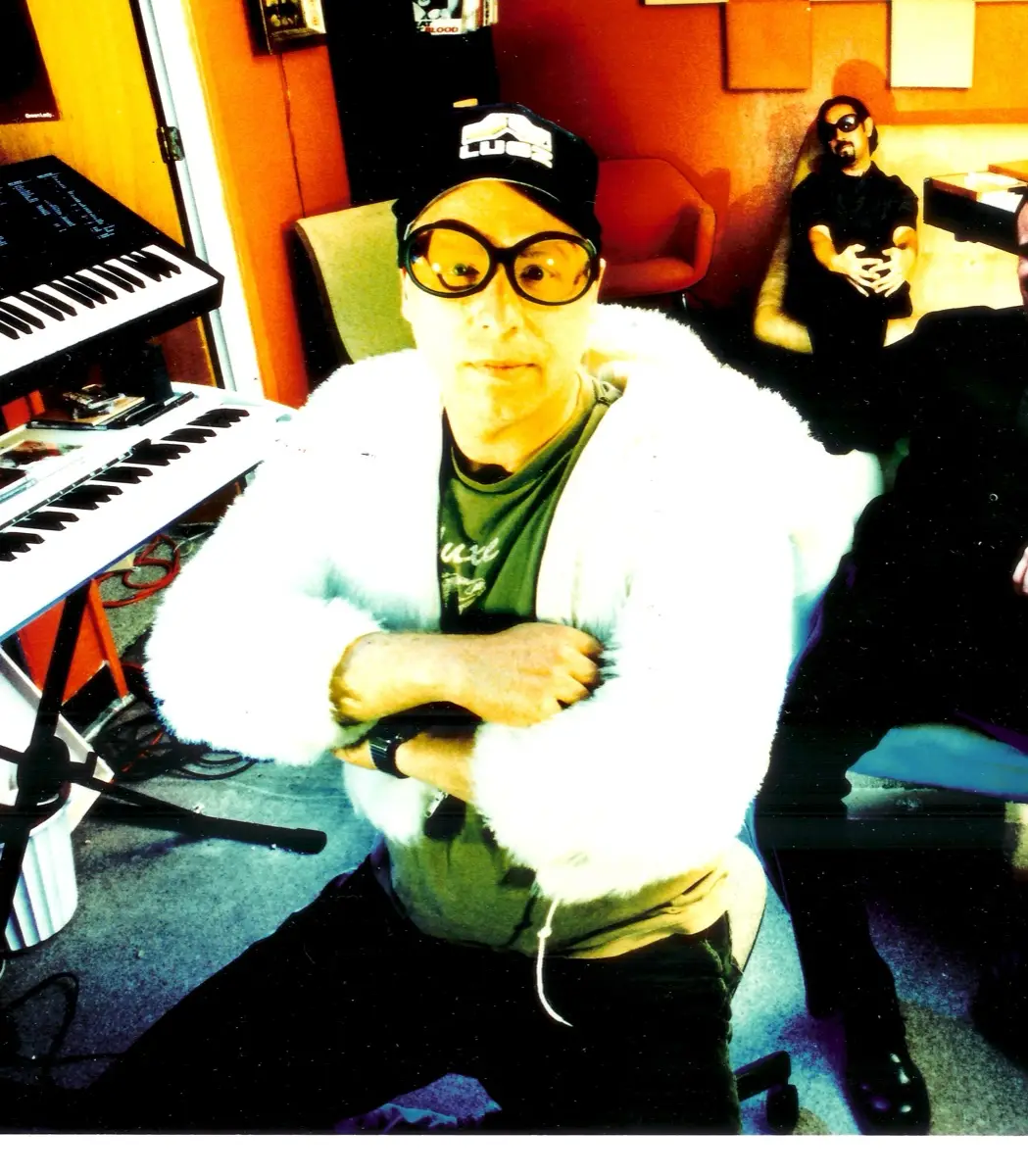 Jim Waters in a white fur jacket, oversized yellow sunglasses and a baseball cap, arms crossed, next to a keyboard.