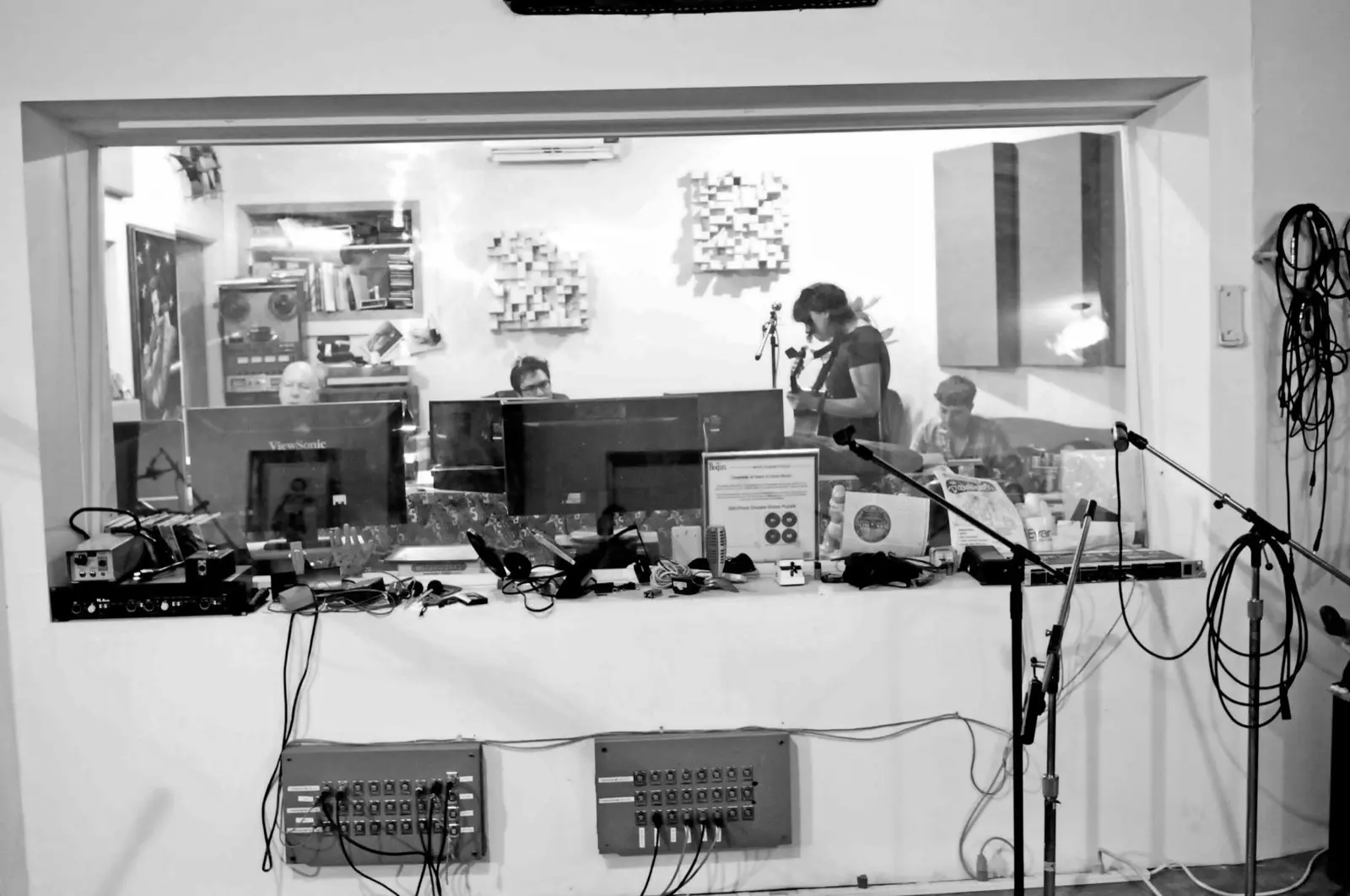Black and white photo from the live room looking into the control room window, with a guitarist and engineers behind the glass.