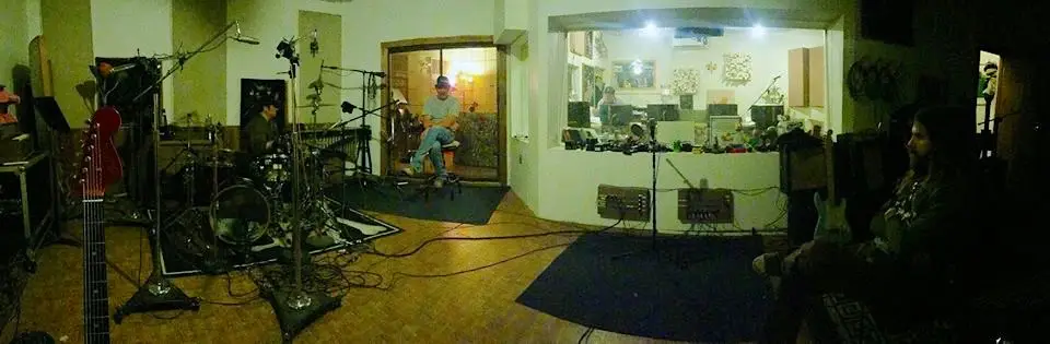 Panorama of the live room with a band set up: drums, guitars, amps and mic stands on rugs, and the control room window behind them.